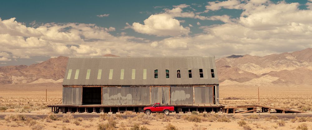 a red truck parked in front of a gray abandoned warehouse in a dry, dry, dry desert. dramatic cumulus clouds above the scene.