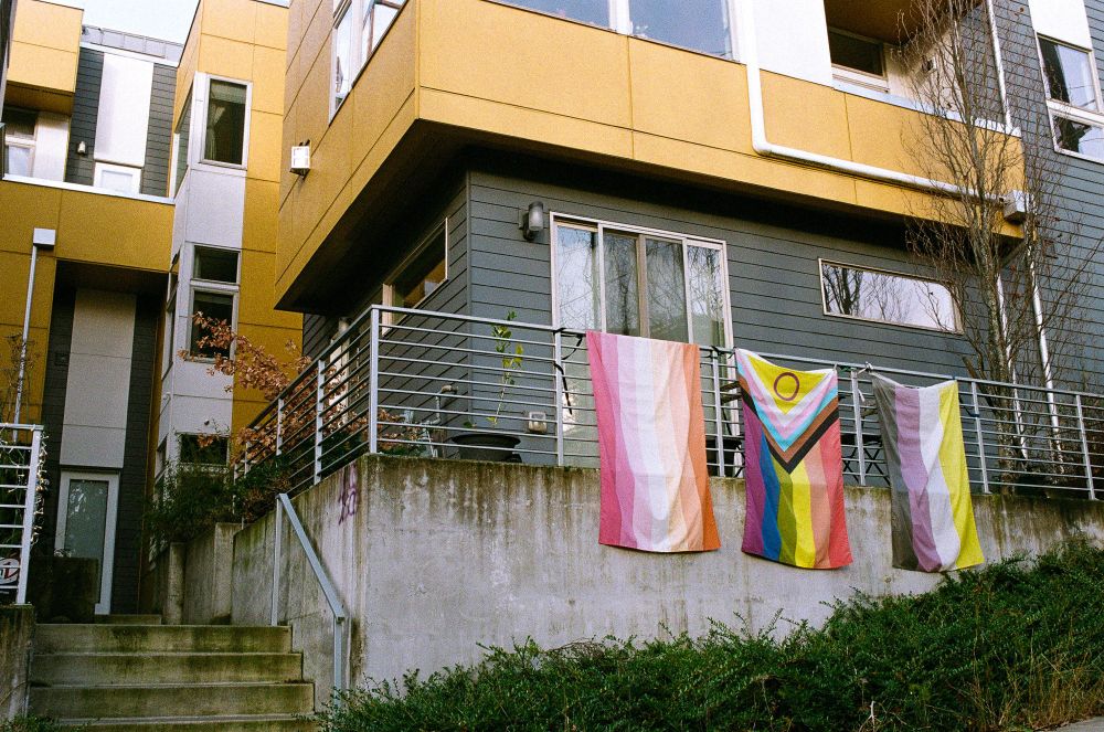 A yellow apartment building with a lesbian, intersex inclusive pride, and NB flag on a balcony out front. 