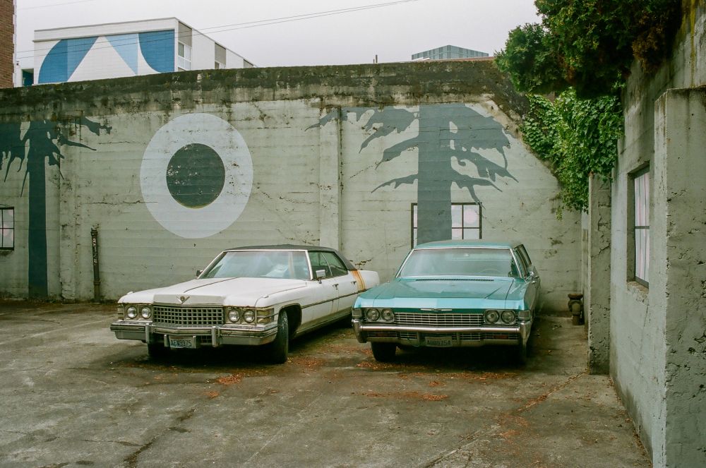 an old cadillac and old impala sit in the back of a weirdly painted concrete-walled parking lot