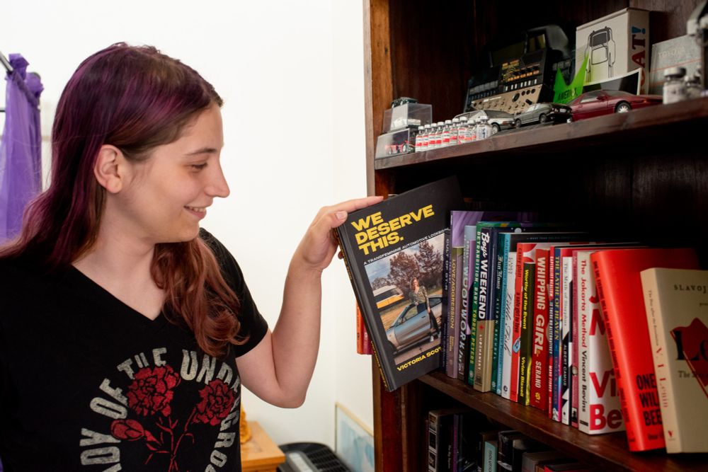 my wife smiling as she pulls out We Deserve This from a shelf containing books including Lenin's collected writings, The Jakarta Method, Boys Weekend, Whipping Girl, Little Fish, Detransition Baby, The Lathe of Heaven, etc. The shelf above has like nine thousand empty estrogen bottles surrounded by little synthesizers and car models