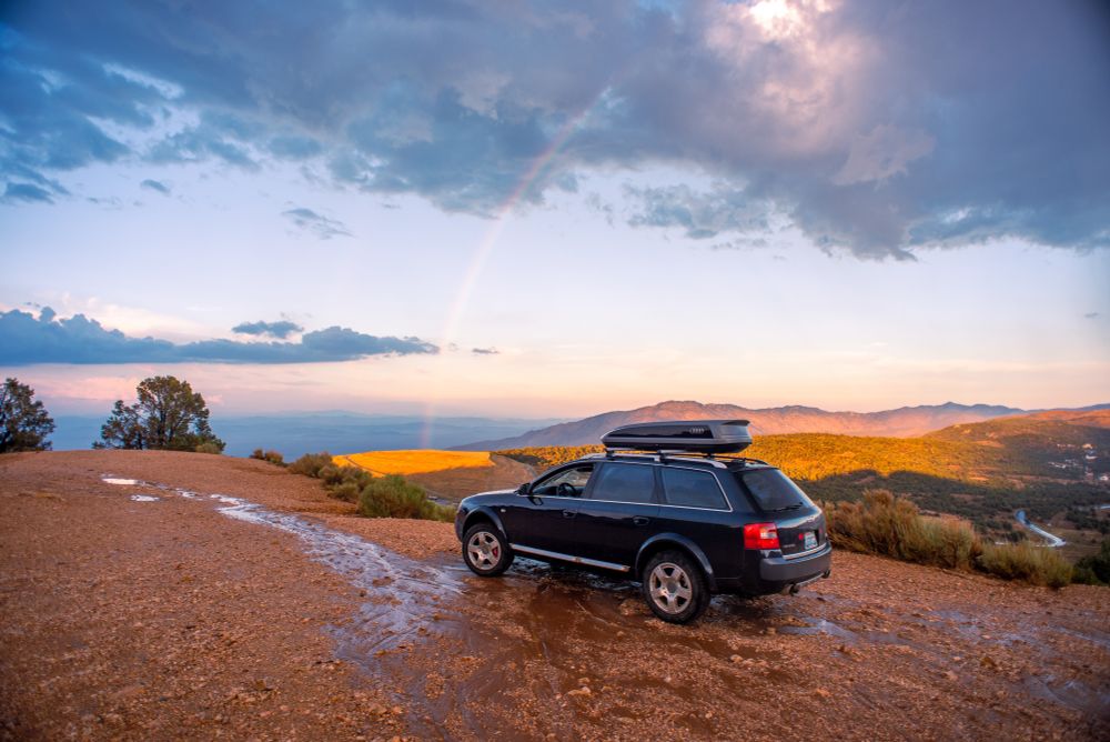 An Audi Allroad with a roof rack sits on a muddy dirt road. Behind it, a rainbow stretches over golden mountains on a pastel sky. 