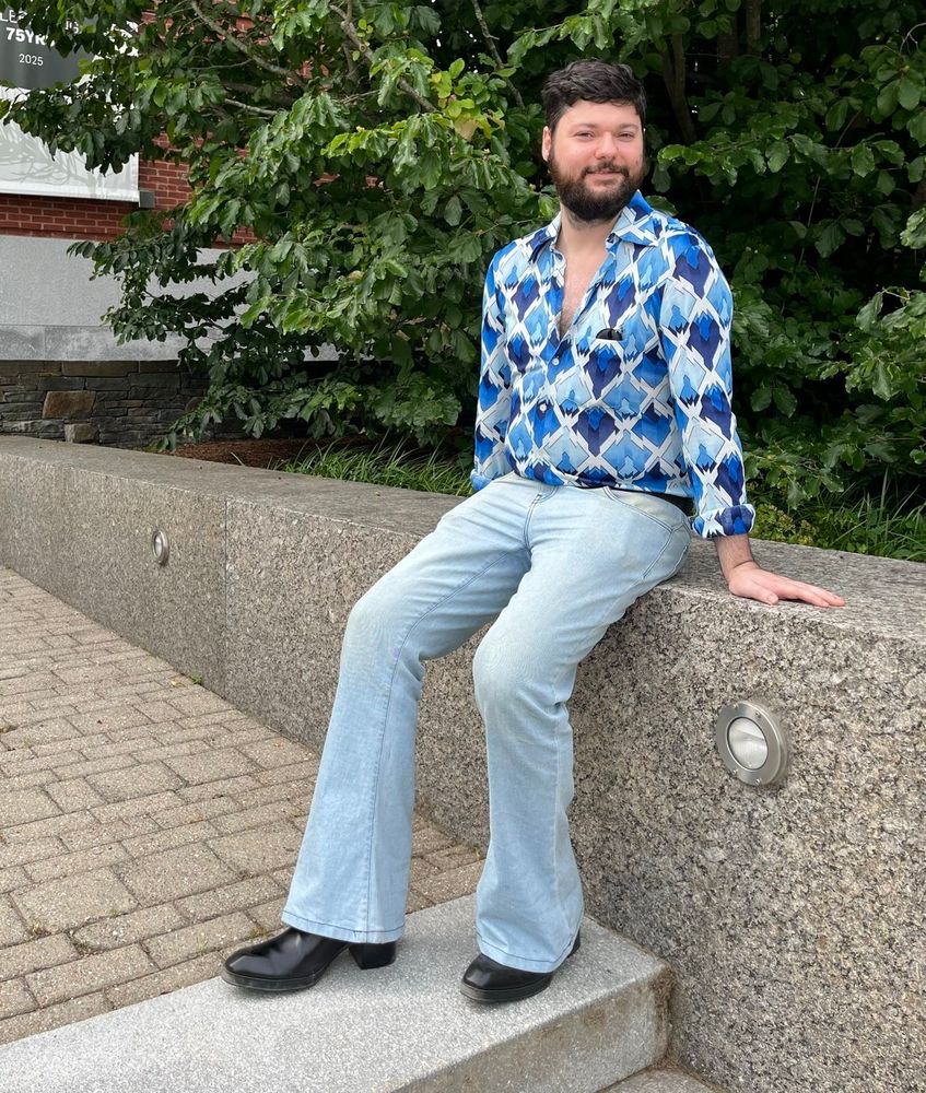 A man sitting on a granite wall in a blue disco shirt with a geometric pattern, lightwash flared jeans, and black high heels.