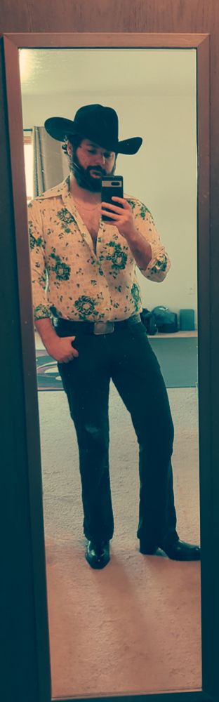 A man wearing an outfit consisting of a black cowboy hat and black heeled boots, dark green cord flares, and a white disco shirt with a green floral pattern