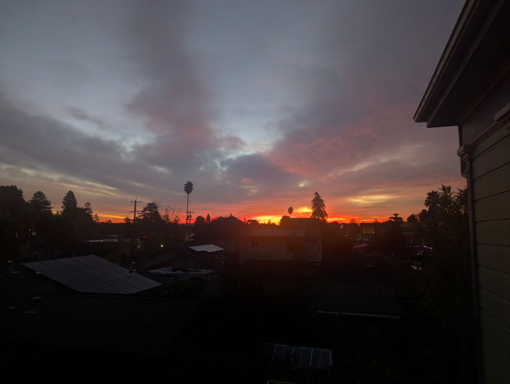 First minutes of sunrise with a hot glowing orange glow the lower part of the horizon, with gray and blue plain clouds above. Picture taken from second floor of a Victorian, building elements visible at edge of picture. 