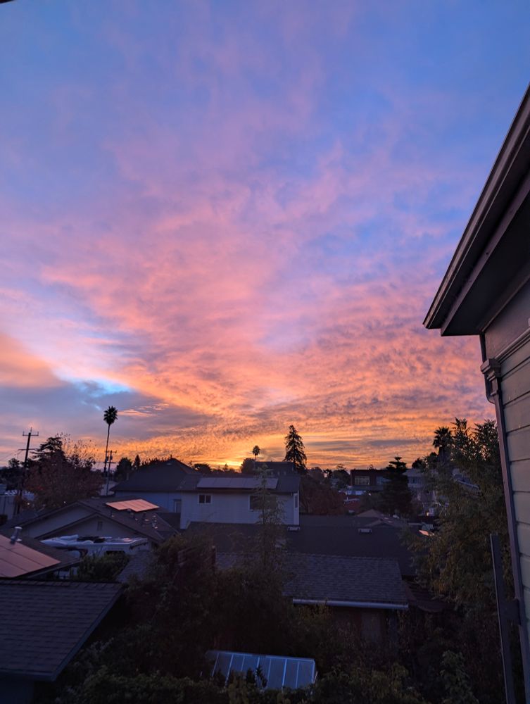 Spread of sunrise with a flowing orange glow coming up from the horizon, with gray and blue plain clouds above. Picture taken from the second floor of a Victorian, building elements visible at the edge of the picture. 