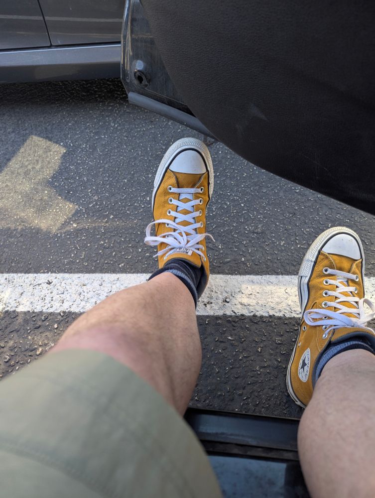 Point of view shot of two legs in a pair of shorts. Sticking out of a car door and out on the pavement. The left foot is resting on a parking stripe and tilted to make an l at the end of that straight leg. The right foot is canted a little outward so that you can see the star of the Converse logo. The focus on the image is the bright yellow-orange padded-ankle Chuck Taylor high top sneakers. 
