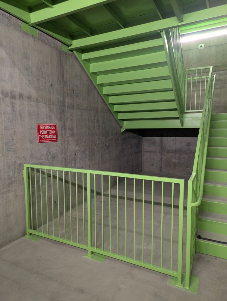 The image is of the bottom of an emergency exit staircase, the stairs painted bright green and the walls bare grey concrete except for a red sign with white letters that reads, 