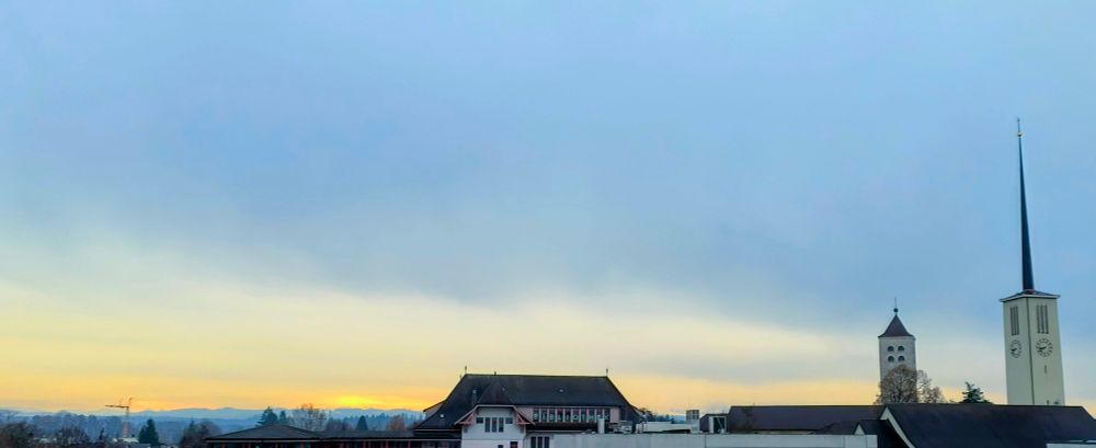 Rechts 2 Kirchentürme, mitte Schulhausdach, links Sonne unter Wolkendecke scheinend.