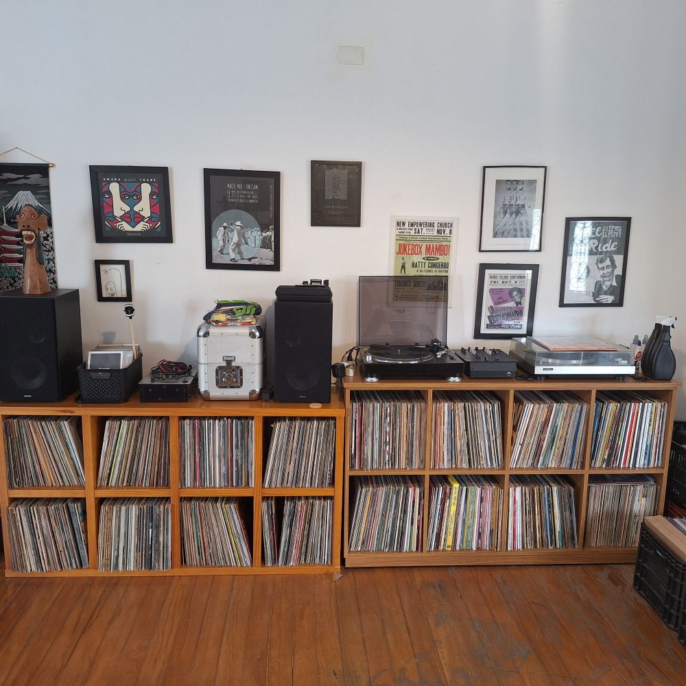 Imagem de uma sala com 2 estantes horizontais cheias de discos de vinil, em cima de uma tem 2 toca discos e um mixer e em cima da outra tem 2 caixas de som, uma caixa de compactos e um case prateado de discos. 
Na parede diversos quadros relacionados a musica e shows. 

