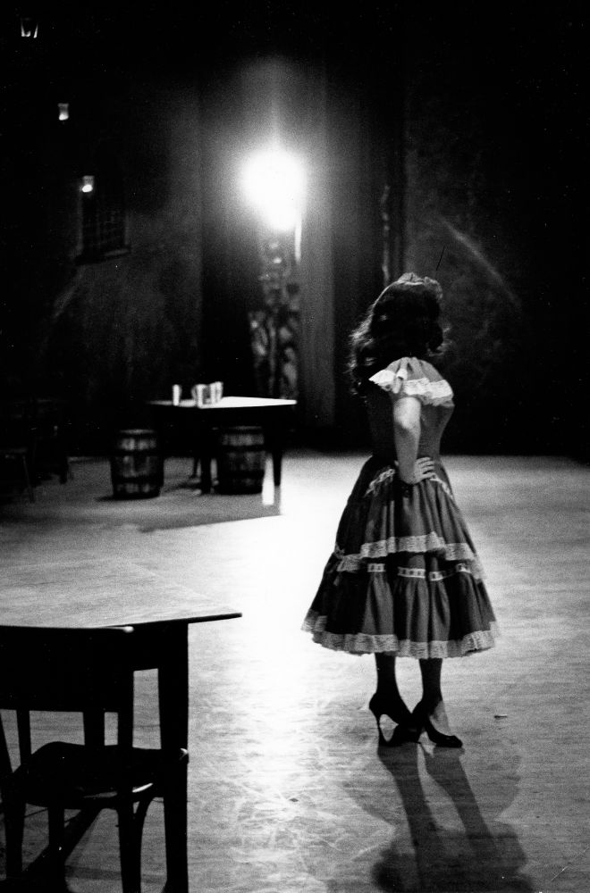 A black and white photo shows a woman standing on a stage. She stands facing right, with her face completely obscured by her dark, long hair. She wears a calf-length dress with loose, short, lace-trimmed sleeves and a skirt of two tiers of lace trimmed layers, and dark, close-toed high heels. She rests her hand on her hip. In front of her is a square wooden table with one chair, and behind her in the distance, another square table with three drinking cups on it and two barrels as chairs. Stage lights pour out from above the table, and two backstage curtains hang on either side of the light. 