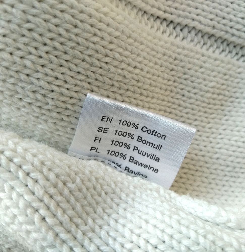 Details of a white stockinette knitted fabric, with a tag stating that it's 100% cotton, in English, Swedish, Finnish and Polish