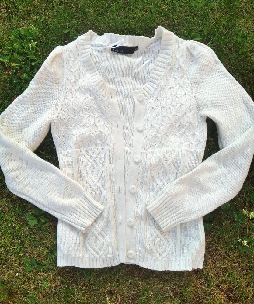 A white cardigan sweater with knitted texture and cables, arranged on partially dry grass