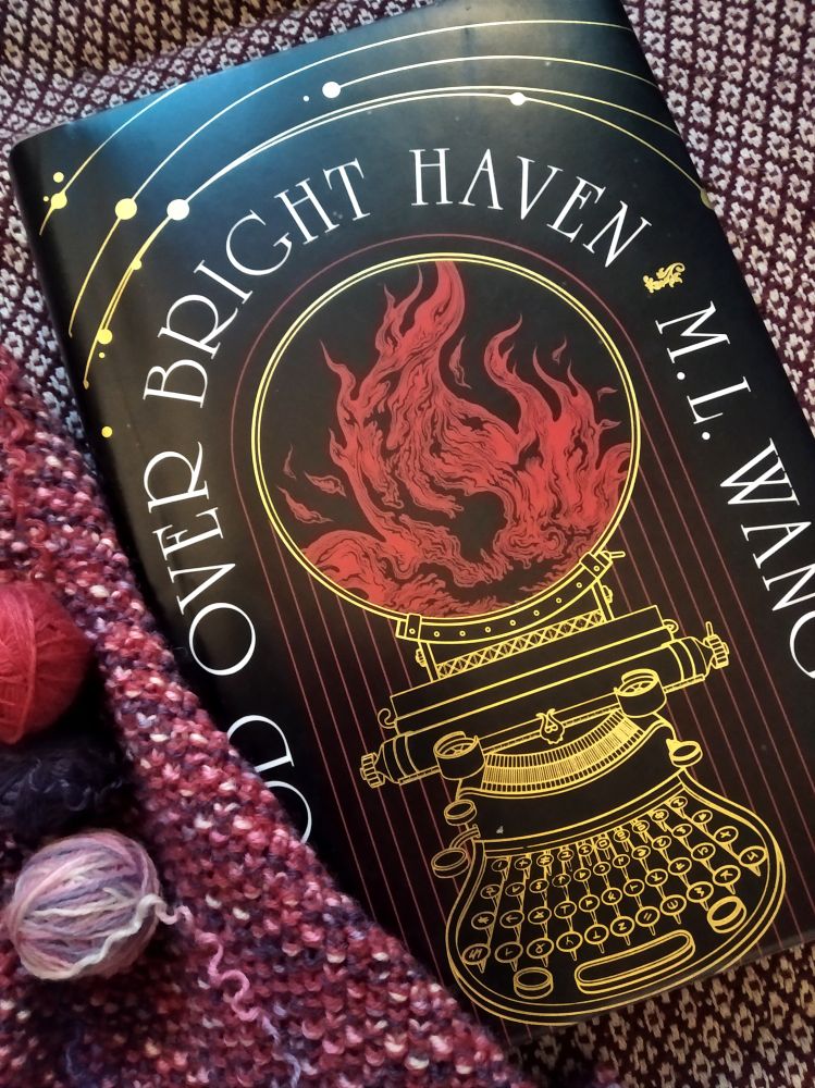 The cover of "Blood over bright haven" book by  M. L. Wang, with a typewriter-like apparatus on the cover etched in gold on black background, framed by a knitted fabric and yarn balls in pink and purple shades