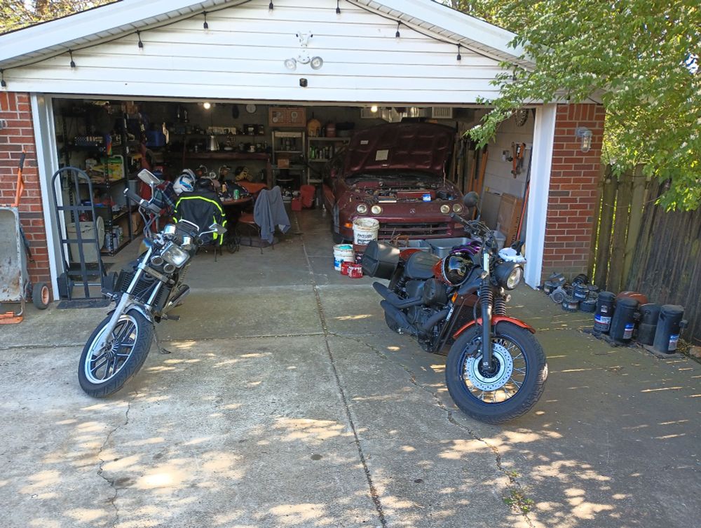 Daytime garage scene with two motorcycles and a red Acura Integra. The black-and-orange honda shadow phantom is parked nearby with the honda Magna to its left. The garage is open and sunny with tools, oil containers, and a tree casting shadows.
