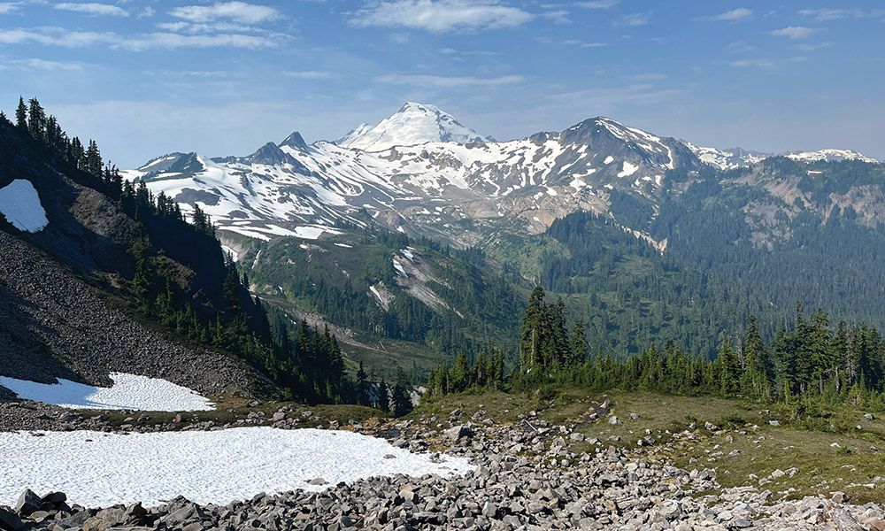 Panoramic view of Mount Baker near a fire lookout tower, with snow-capped peaks and surrounding lush green forests under a clear blue sky.
