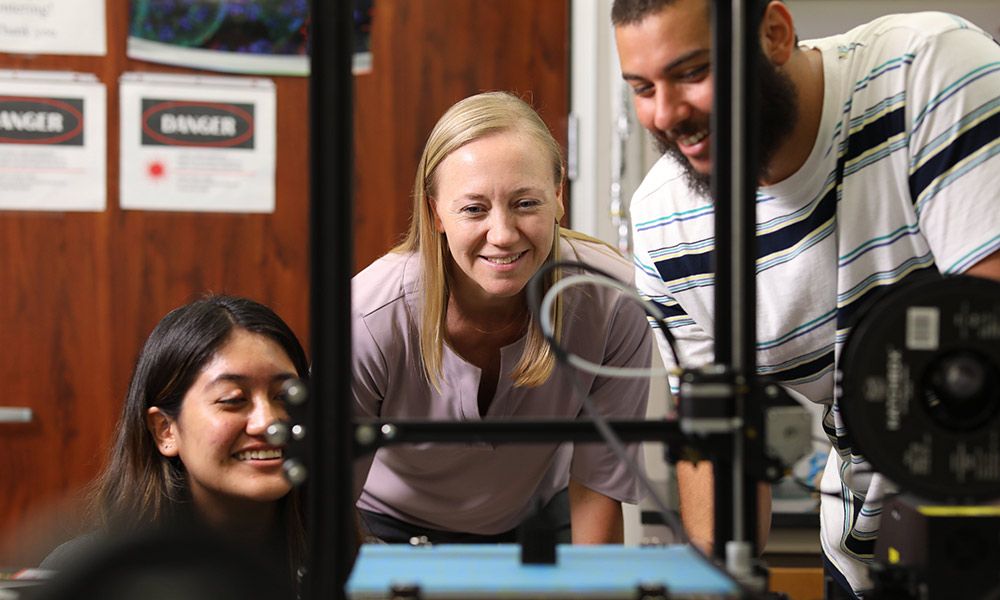 Three people are smiling as they look at lab equipment. 