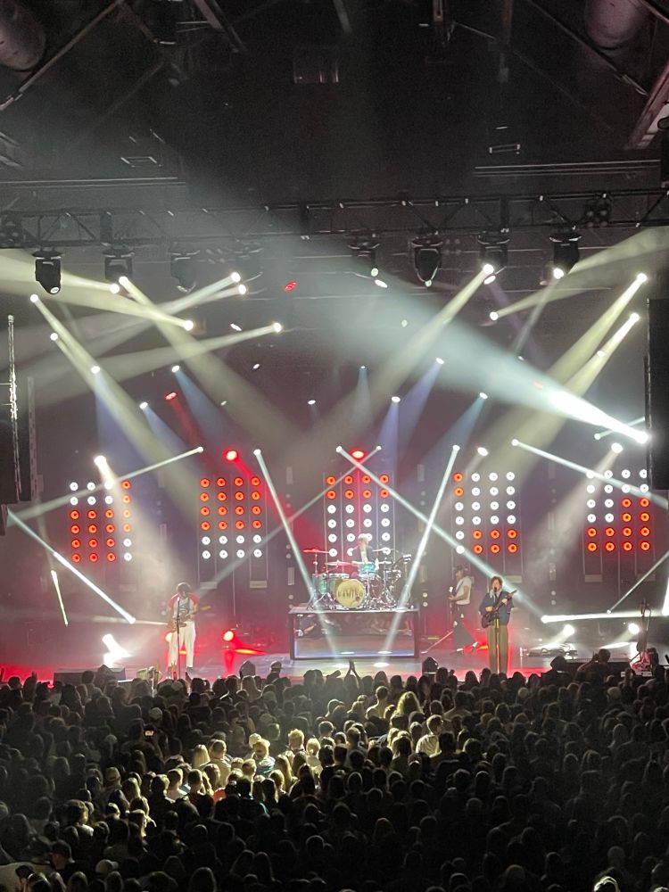 the band CAMINO performing on stage in front of an audience. There are white lights coming down from the ceiling and red and white lights at the back of the stage 