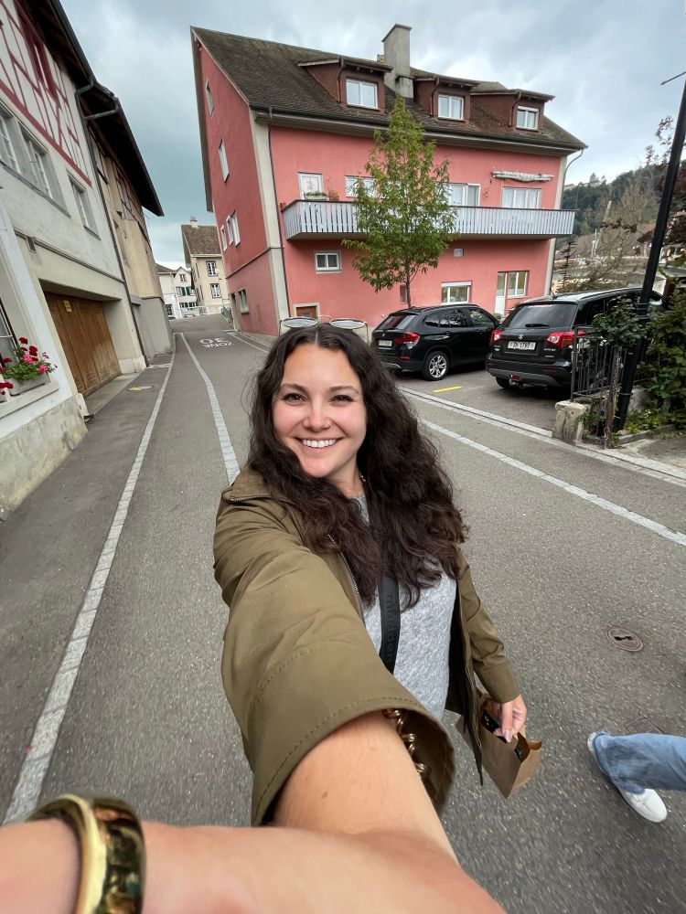 0.5 selfie of me walking along a street in switzerland! I’m wearing an olive green rain coat, a gray shirt and my hair is brown and wavy/curly & I’m smiling! 