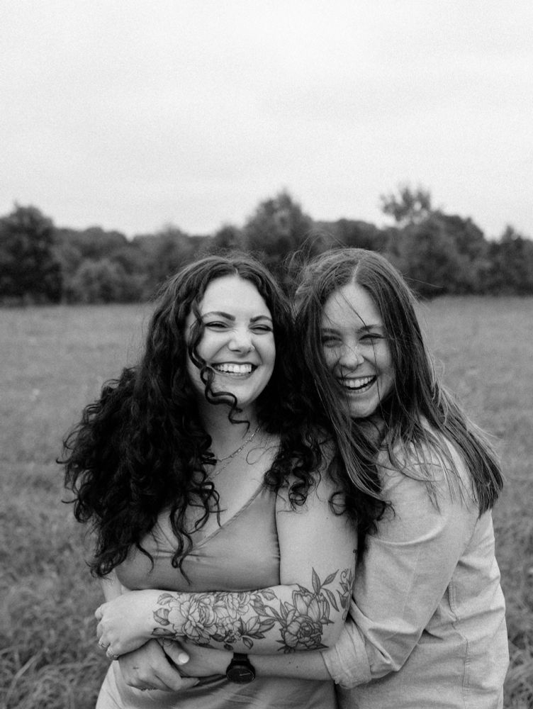 black and white picture of my wife Andi and me. we’re both smiling. I am on the left and I have dark brown curly hair with floral tattoos on my left arm. Andi is on the right and she has straight brown hair and is wearing a long sleeved button down, with the sleeves rolled up on her forearm 