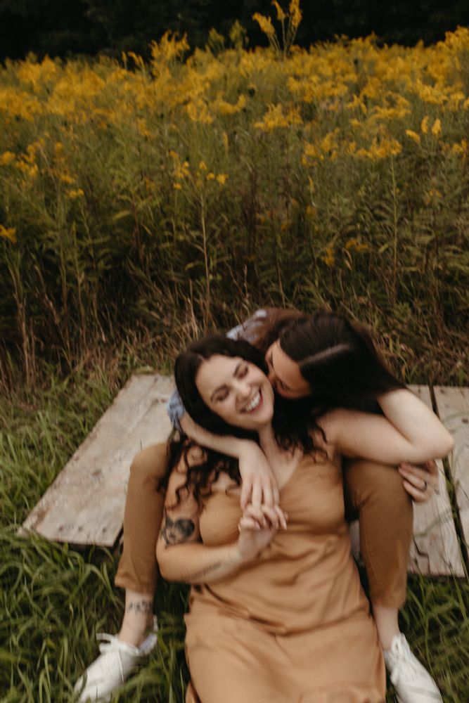 me and my wife (fiancé at the time). I’m sitting on the ground in front of her, in between her legs and she’s kissing the side of my head while I’m smiling. We’re holding hands with our right hands and my left hand is holding Andi’s head. we are sitting in a field 