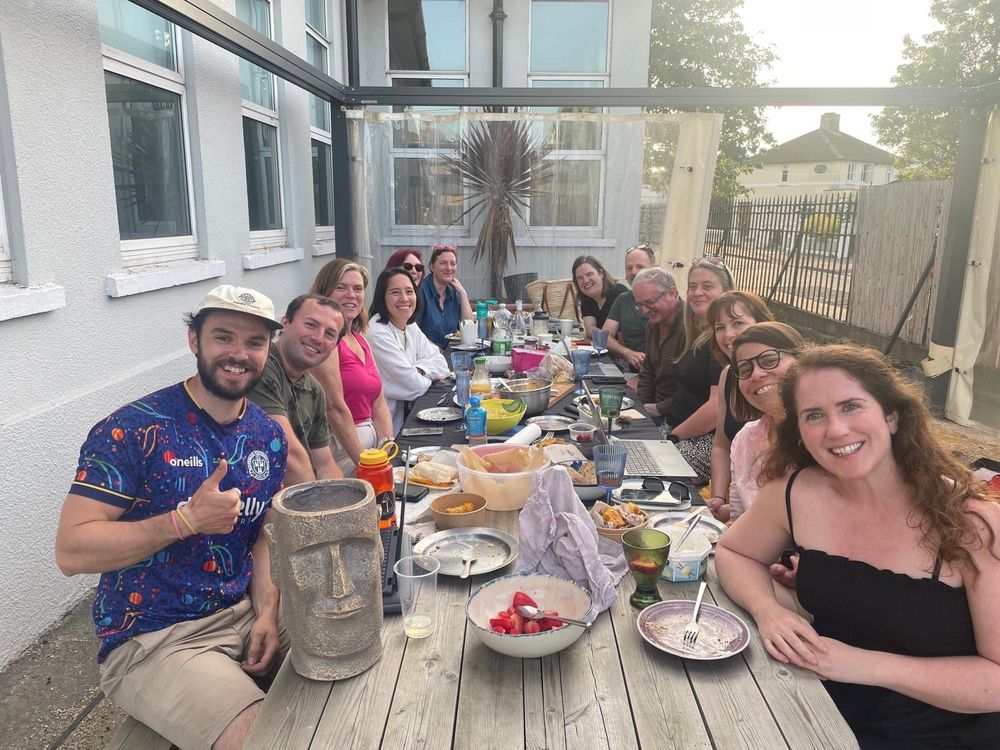 The Connecting Cabra committee - 12 smiling people sitting along a large picnic bench with food laid out on the table. 