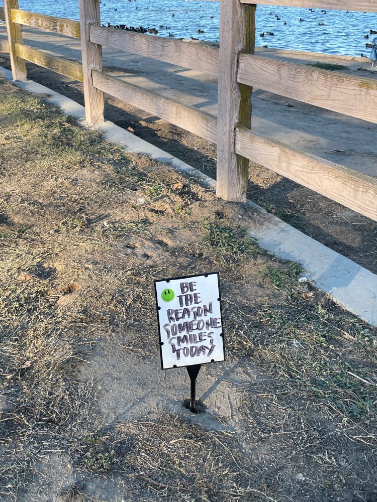 A sign on which someone has handwritten “Be the reason someone smiles today.” The sign is propped up in a patch of dirt and grass next to a lake with ducks.