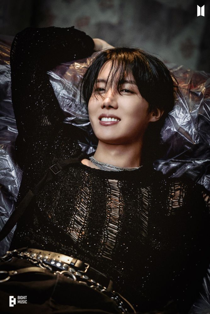 j-hope of BTS, wearing a black mesh sweater that teasingly reveals his bare chest beneath it, reclining on a plastic covered couch. He is smiling his heart-shaped smile beautifully, but also staring into the camera with a glint in his eyes that says, “I know you want me.” Yes sir, we do!!!