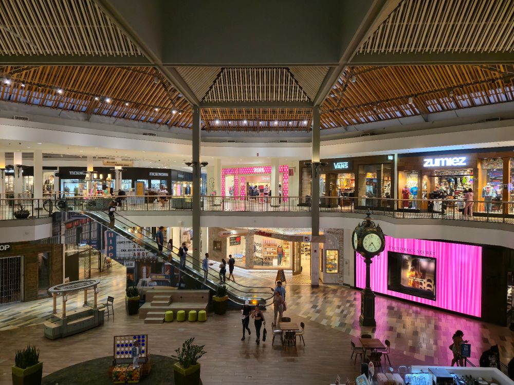 Overlooking a central court of a bi level shopping mall. An escalator is in the background, a clock tower to the right. Wood slatted ceiling fixtures provide ambience and a comforting environment. 