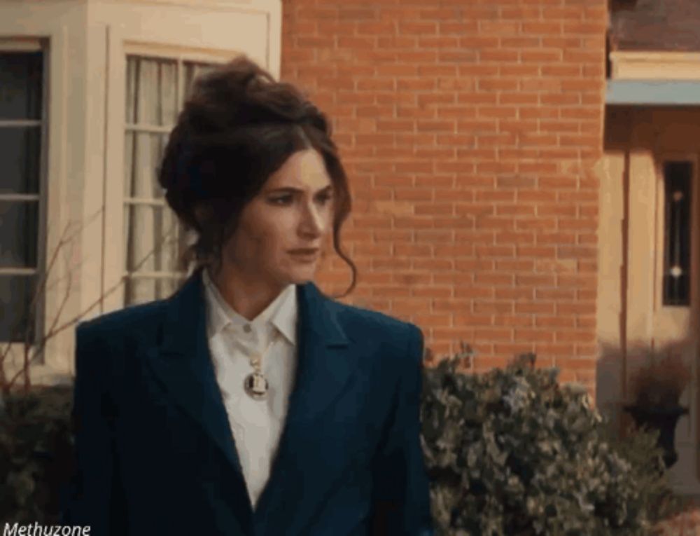 a woman in a blue suit and white shirt is standing in front of a brick house .