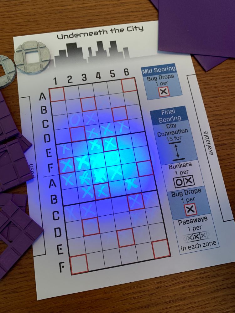 A photo of my game Spy Moles. There's a sheet of paper with a grid on it, and a blacklight shining on the paper reveals markings made by the player with an invisible ink pen. Surrounding the paper are cards and stencils