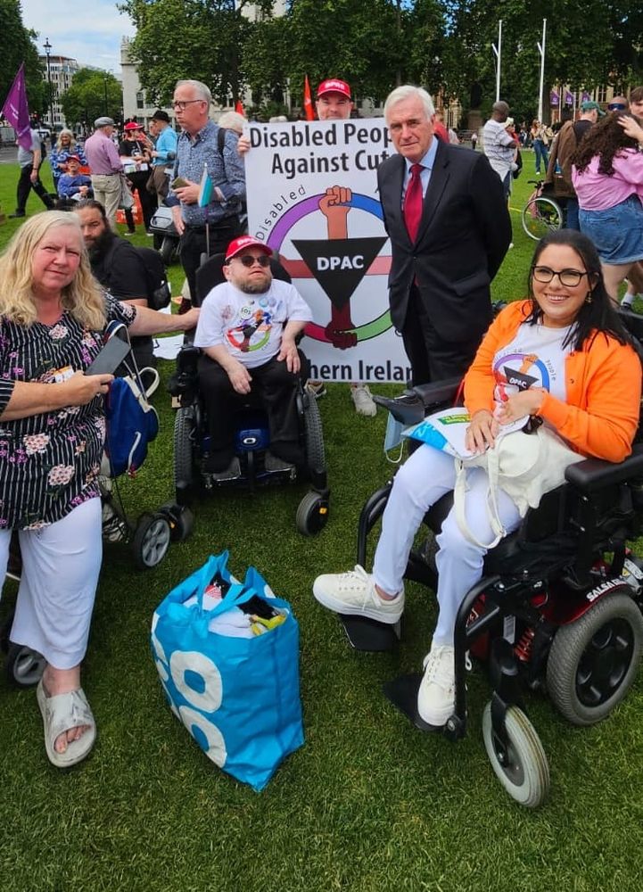 Group of people, including individuals in wheelchairs, stand outdoors on Parliament Square London, outside Westminster. WIth them is Labour MP John McDonnell.

They are in front of a sign that reads "Disabled People Against Cuts Northern Ireland.