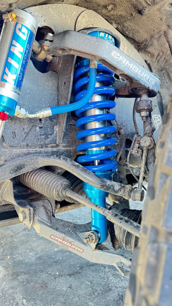 A picture of the steering rack of a 2017 Toyota Taocoma. With a 2.5 Coil Over Shock with Blue Springs and a King Off-Road shock remote reservoir with red adjuster knob. Bolted into a Camburg Kinetic Upper Control Arm and Camburg Trail Series Long(ish) travel Lower Control Arm, Half Axles, Tie Rod Link, and Sway Bar are also visible 