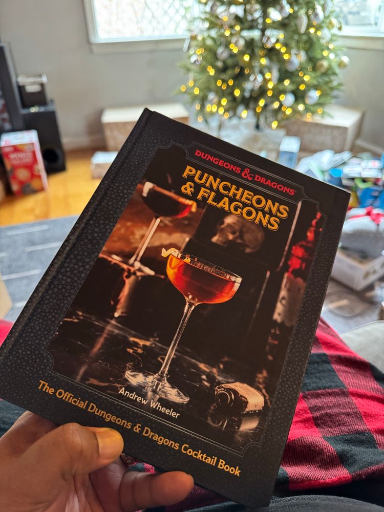Hand holding ornate book with fancy cocktails on cover. Titled Puncheons & Flagons. The official Dungeons & Dragons Cocktail Guide 
