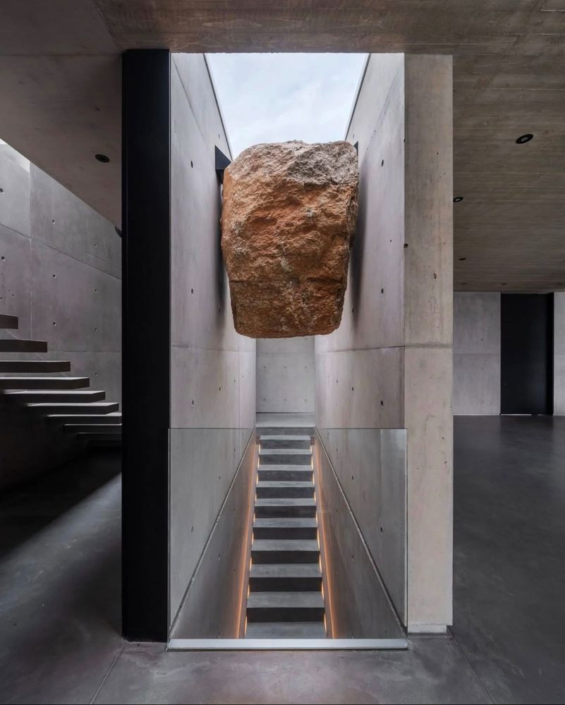 A large rock is elevated above a stairwell - seemingly wedged between two wall - in a contemporary concrete home with no visible furniture.