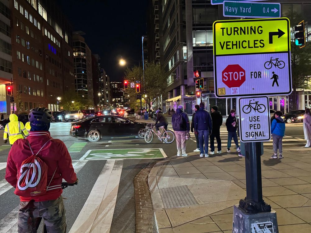 M and 1st Street SE, outside Nats park. A cyclist with a Washington Nationals drawstring bag is stopped next to a sign with a ‘The War on Cars’ sticker on it.