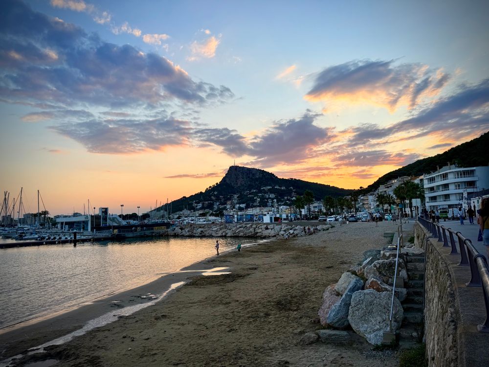 Sunset at L’Estartit marina – A soft evening light over L’Estartit’s marina and beach promenade. The bay is calm, sailboats are moored, and clouds scatter a pink-orange glow across the sky as the sun dips behind the hills.