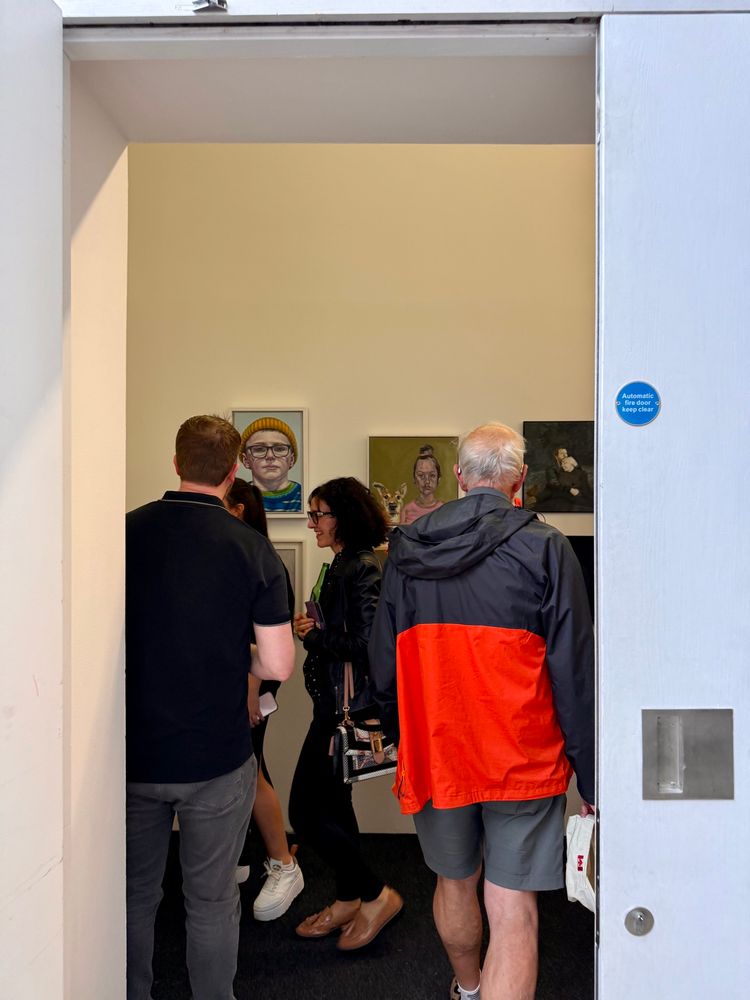 A small group of people gather inside a narrow gallery space, seen through a large open door. Three contemporary portraits hang on a beige wall in front of them, featuring expressive depictions of people. One visitor wears a bright red and black jacket, adding a pop of colour to the neutral surroundings. The setting is lively and intimate, suggesting engaged conversation about the artworks.