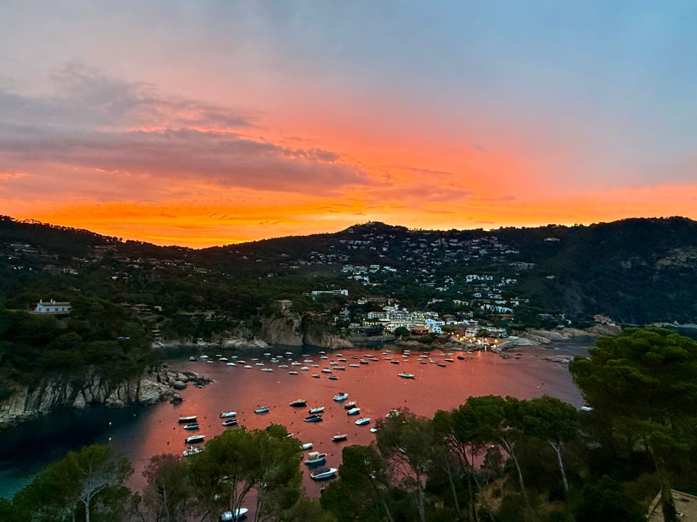 Aiguablava at sunset – A stunning coastal view of Aiguablava, Catalunya, just after sunset. The sky glows orange and pink, with dozens of boats anchored in the calm bay below. Hills dotted with houses rise in the background.