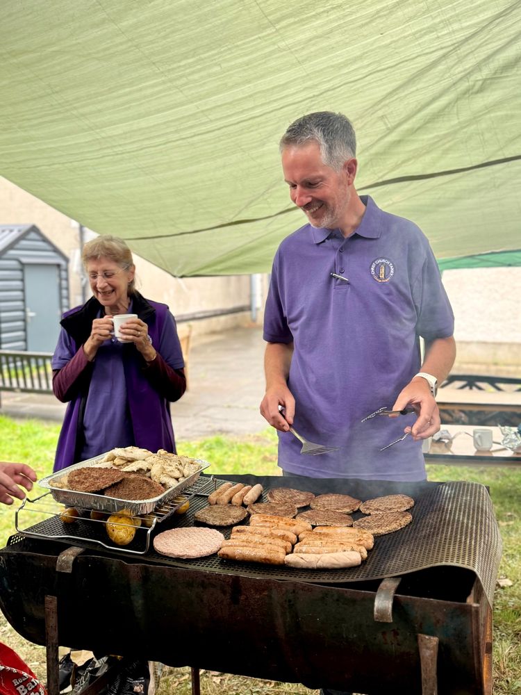 A man in a purple bellringing polo shirt tends to a sizzling BBQ filled with sausages, burgers, and chicken. Another bellringer, an older woman, stands nearby holding a mug and laughing. They’re under a green tarp, keeping dry from light rain.