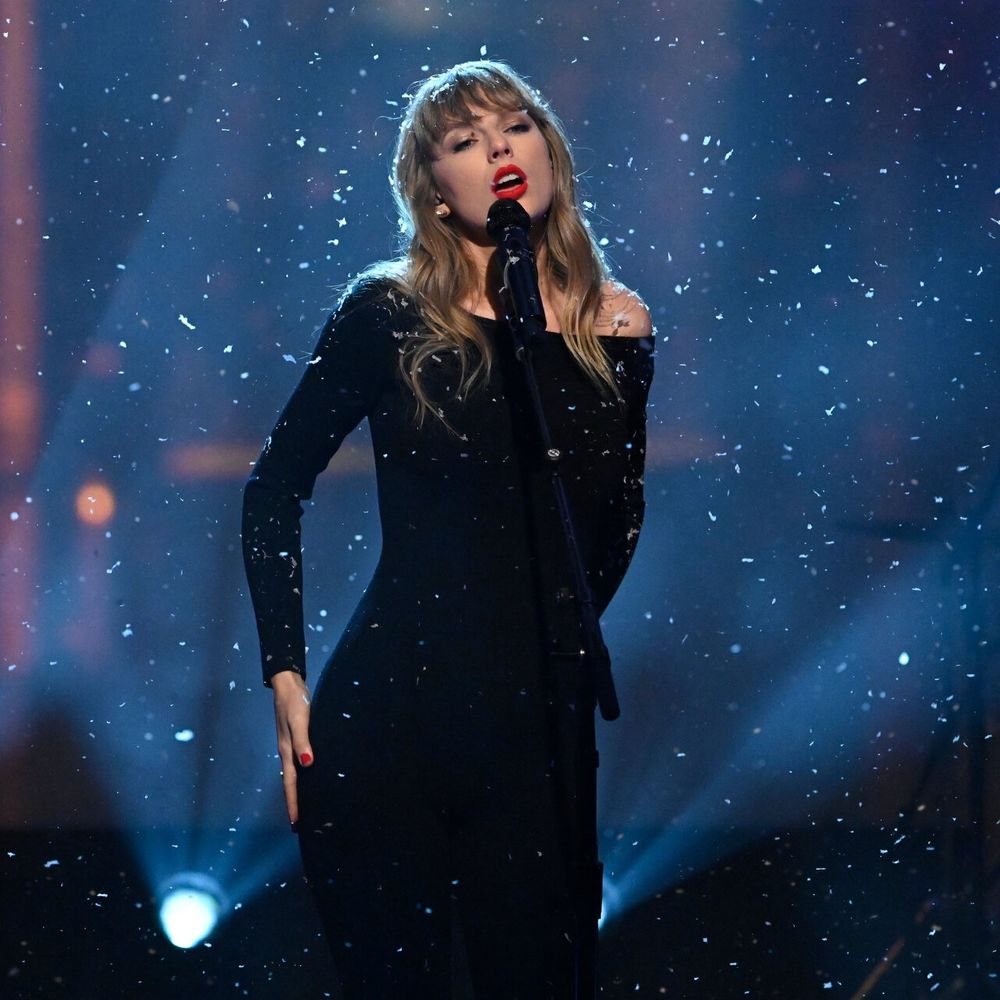 Taylor Swift dressed in all black with red lipstick with fake snow falling on her. 