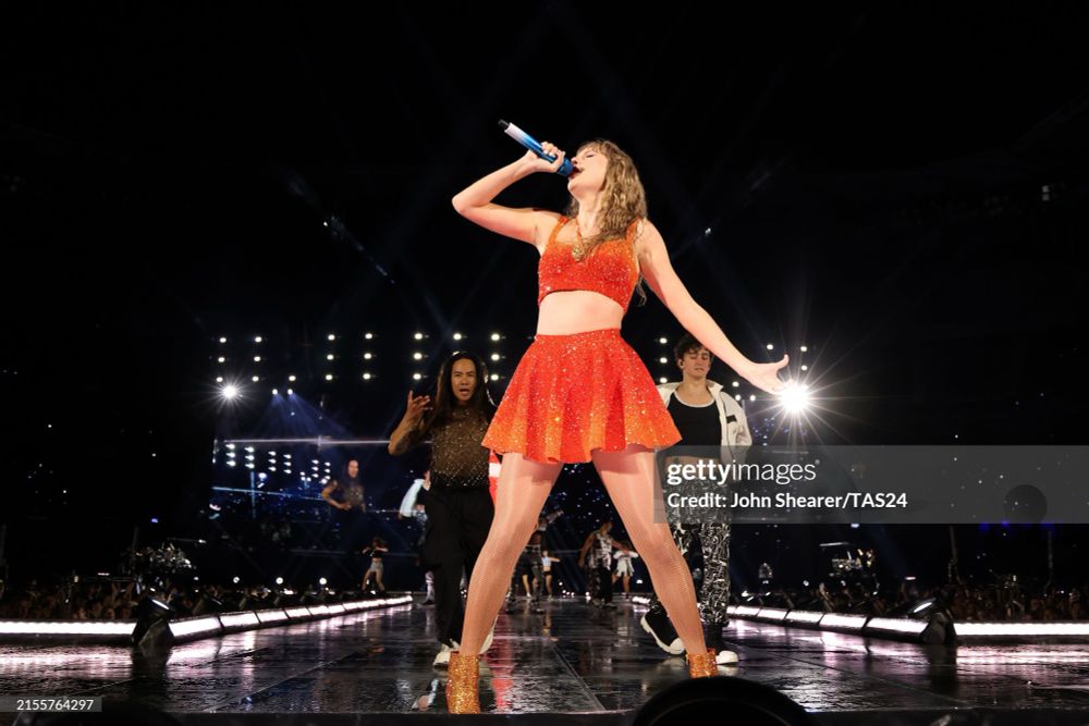 Taylor wearing an orange sparkly top and skirt and singing. 