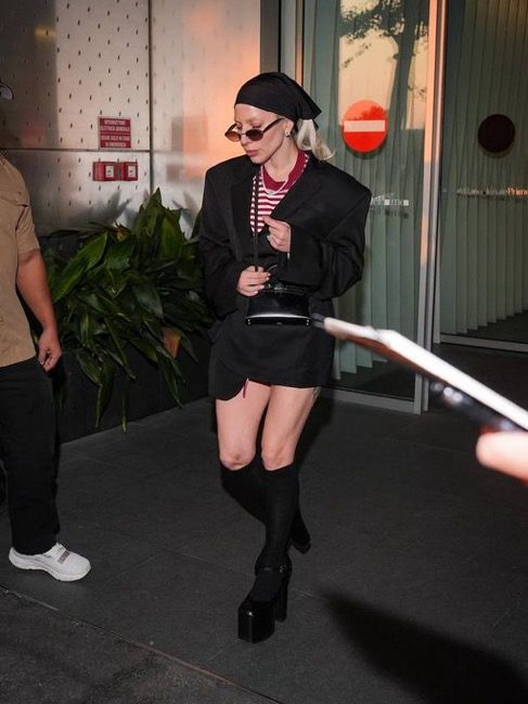 Lady Gaga wearing a red and white striped top with an oversized black coat and black knee high socks with heels. Her hair is currently blonde. 