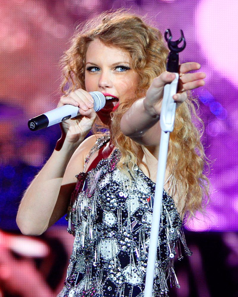 Taylor Swift, singing into a white Mic wearing a black and silver dress. 