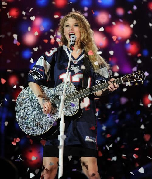 Taylor Swift performing with a bedazzled silver guitar and wearing a patriot number 13 jersey. 