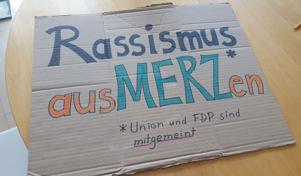 Demoschild mit der Aufschrift "Demokratie aus-merz-en". Merz ist mit einem Sternchen markiert, darunter steht mit Sternchen "Union und FDP sind mitgemeint"