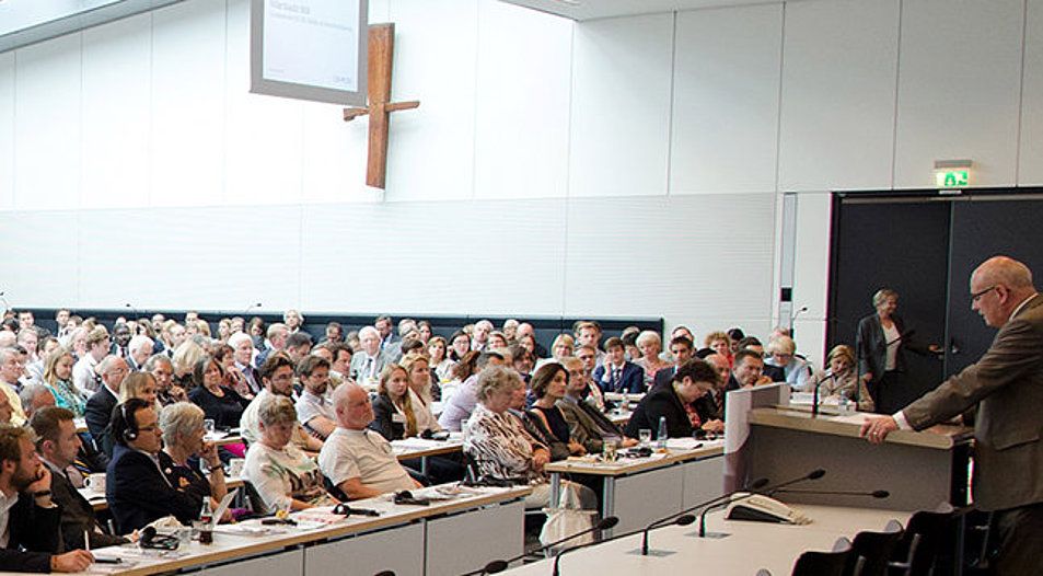 Foto eines großen Saals mit mehreren Personen, die einem Redner lauschen, darüber hängt ein Holzkreuz