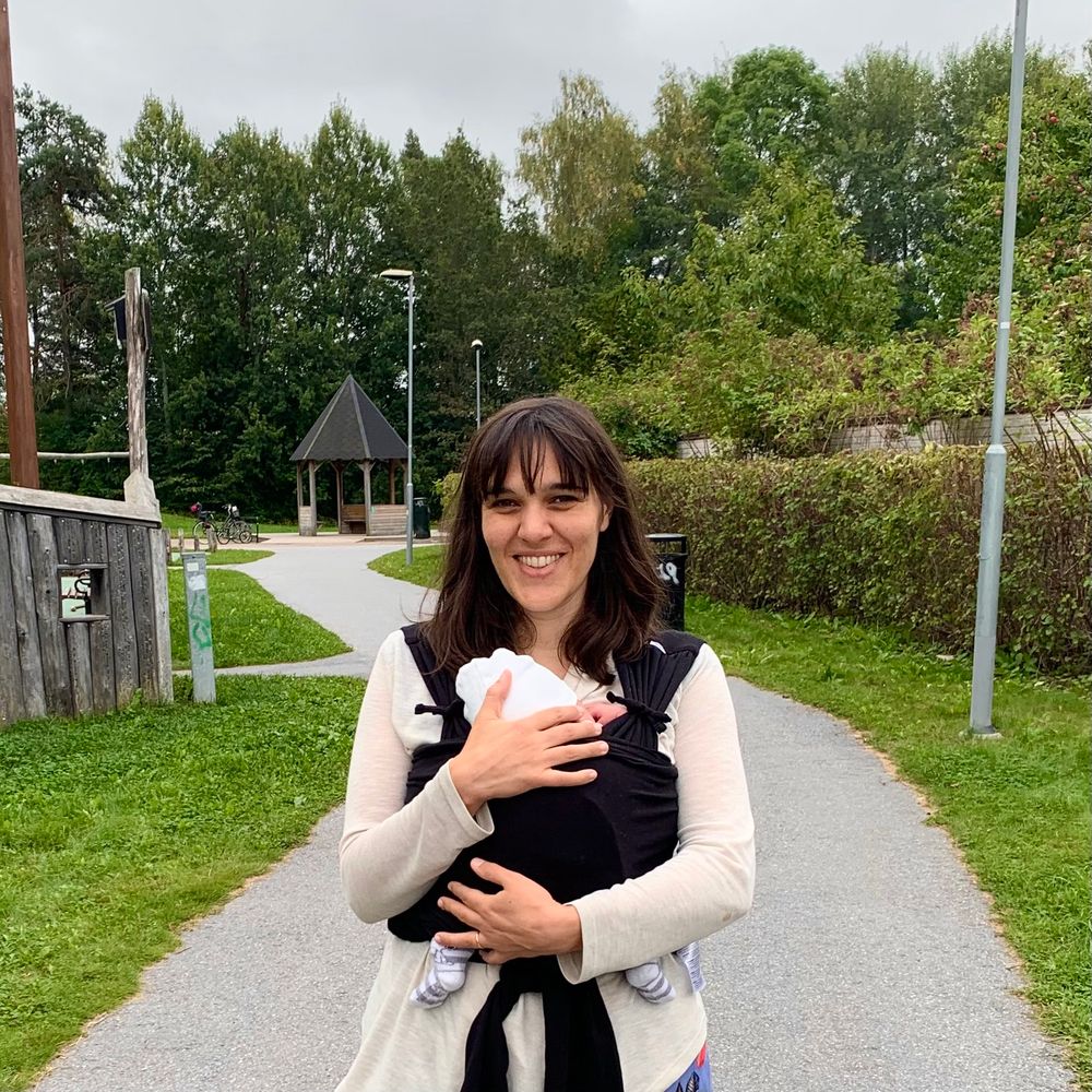 Portrait of me in a park. Im facing the camera smiling. I’m carrying a baby in a wrap in front of my chest. Only a hat and feet of the baby are visible. 