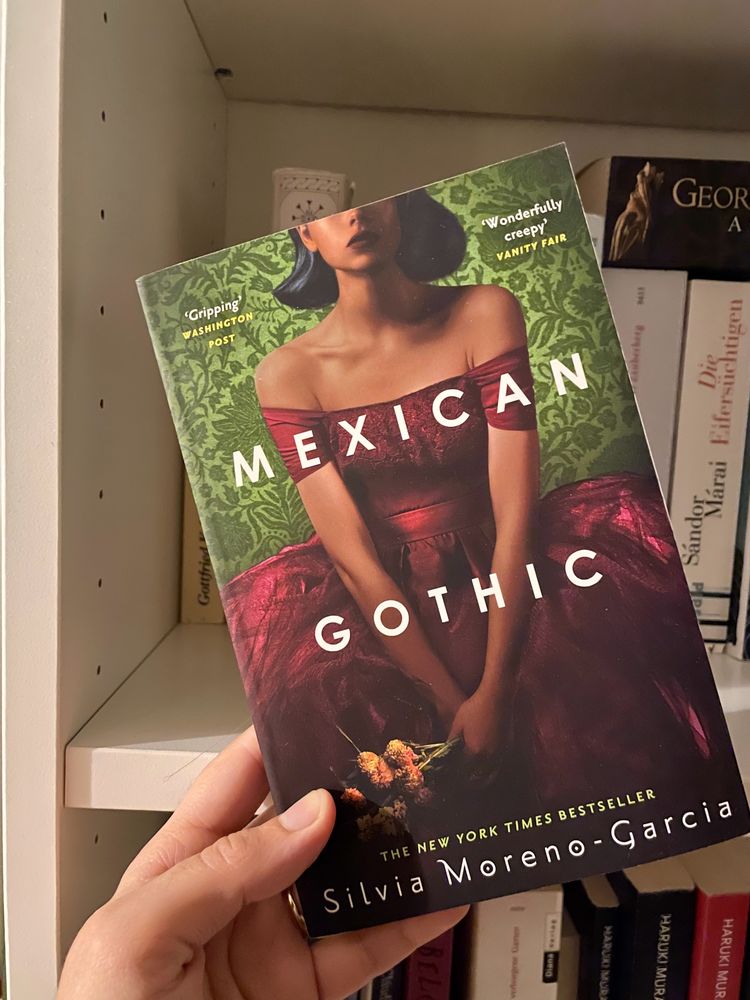 The cover of Mexican Gothic by Silvia Moreno-Garcia showing a black woman in a burgundy gown sitting in front of a green wall. Her face is only half visible.
