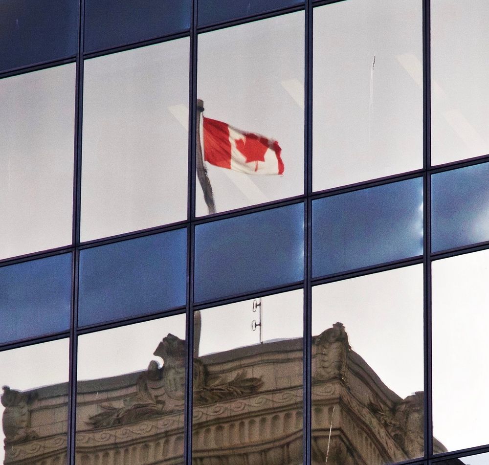 Image of a Canadian flag on a flagpole on top of a stone building, reflected in a glass building window.