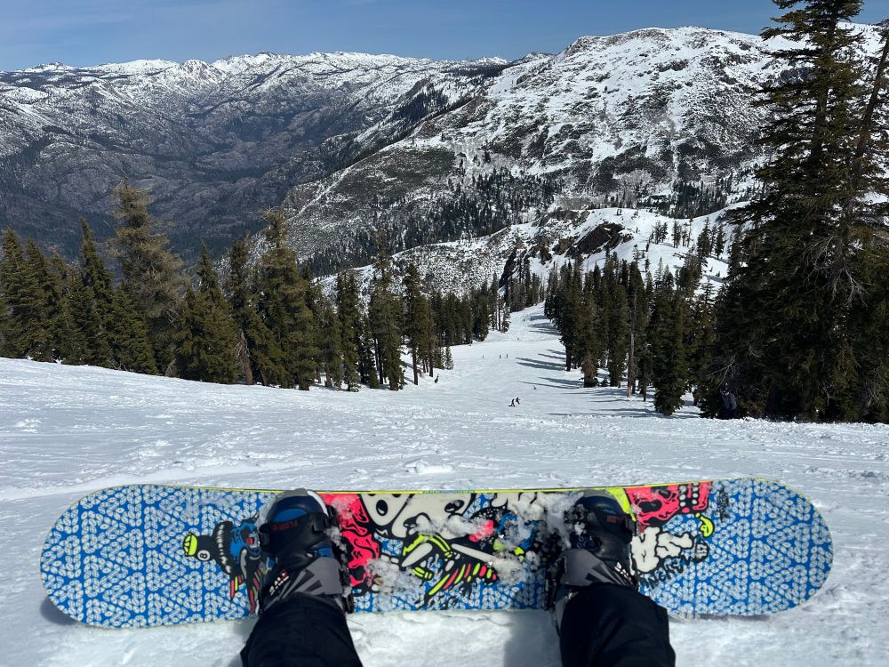 Looking down on a snowy slope at Bear Valley, trees on both sides, with a snowboard in the foreground and snow covered mountains in the background.